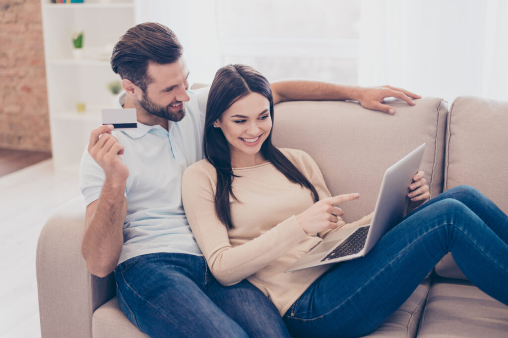 Un hombre y una mujer alegres están haciendo compras por Internet con una laptop y una tarjeta bancaria en casa en una acogedora sala de estar. Pareja aprendiendo a convertir puntos en millas.