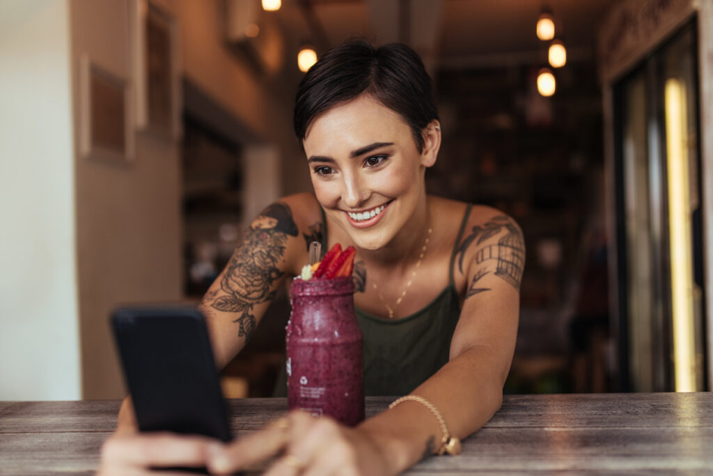 Una mujer con cabello corto y un tatuaje está en una mesa con una taza de acai frente a ella y juguetea con su teléfono celular mientras sonríe. Está feliz de cambiar millas por dinero de manera segura en MercoMillas