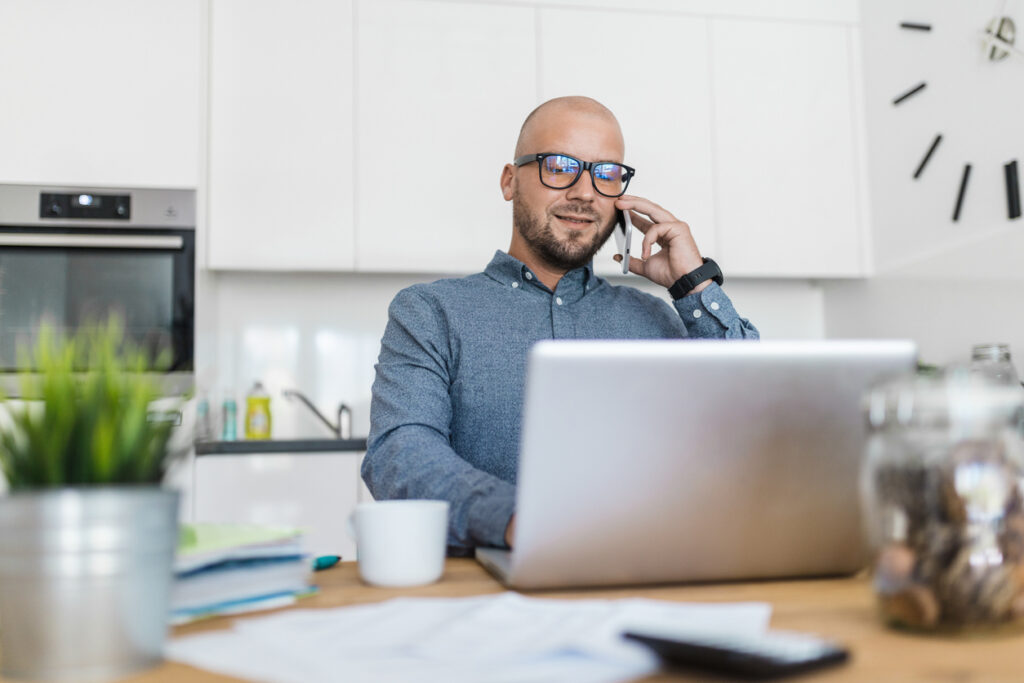 Un hombre calvo con playera gris está sentado en un escritorio frente a su computadora mirando la pantalla mientras conversa por su celular con un asistente de MercoMillas para saber cómo acumular millas