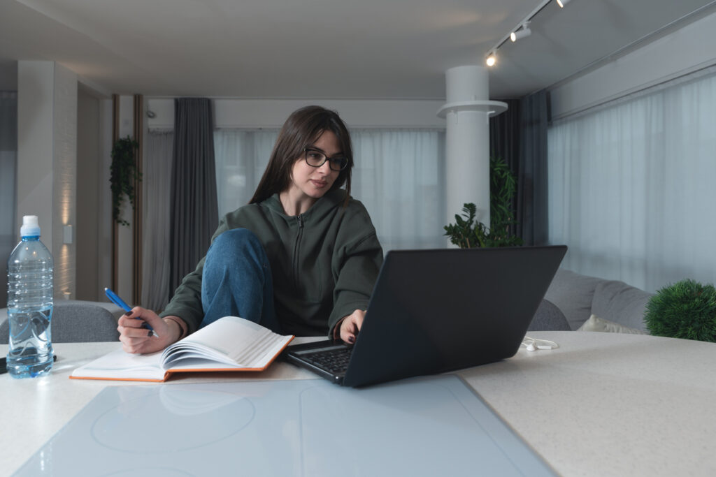 Una mujer blanca con el pelo negro y lacio lleva gafas, una sudadera gris y vaqueros. Ella está sentada en una mesa mirando su computadora y tomando notas en su cuaderno. Está aprendiendo que acumular millas aéreas puede ser rápido y fácil.