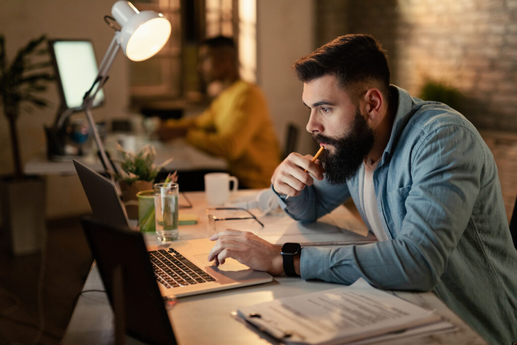 Un hombre blanco con cabello negro y barba negra viste una blusa blanca y una camisa de mezclilla azul claro. Está sentado en un escritorio frente a su computadora, con un lápiz en la boca mientras lee consejos para no cometer errores al vender millas de aéreas