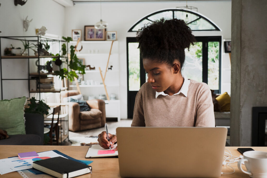 Una mujer negra con un suéter rosa claro y el cabello recogido hacia atrás está sentada en una mesa trabajando en la computadora y escribiendo en un cuaderno. Está anotando los mejores consejos para disfrutar de sus millas aéreas